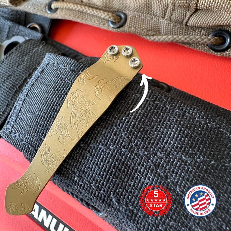 Puede incluir: Un clip dorado para navaja de bolsillo con un dise&ntilde;o de pez koi grabado. El clip est&aacute; sujeto a una funda de nailon negro. La imagen tambi&eacute;n incluye una pegatina de "5 estrellas" y "American Product Made in USA".