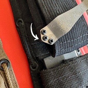 May include: Close-up of a silver metal knife blade with an etched koi fish design. The blade is attached to a black nylon sheath with three black screws. The background includes a red and tan surface.