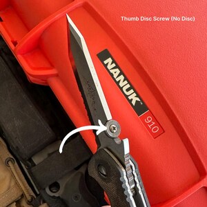 May include: Black folding knife with a serrated blade and a thumb disc screw. The knife is partially open, revealing the blade's edge. The handle is black and textured. The knife is next to a red case with the text "NANUK 910".