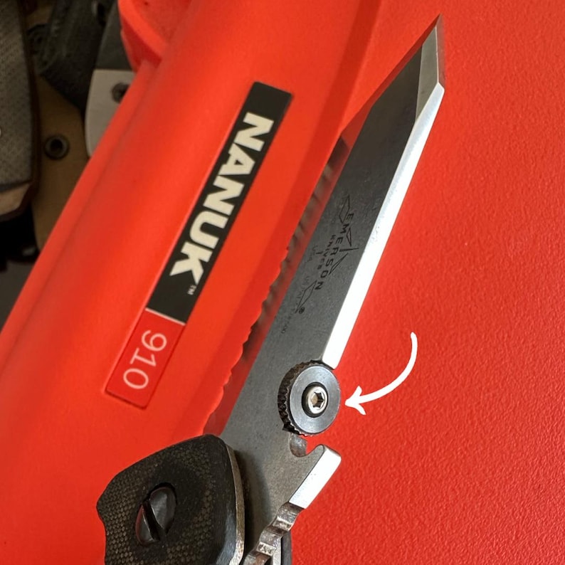 Peut inclure: Gros plan sur un couteau pliant noir et argent&eacute; avec une lame partiellement dentel&eacute;e. Le couteau a un manche noir textur&eacute; et un m&eacute;canisme de pivot argent&eacute;. La lame porte la mention "Emerson Knives". Le couteau est pos&eacute; sur un fond rouge.