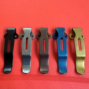 Puede incluir: Cinco clips de bolsillo met&aacute;licos en varios colores: negro, plateado, bronce, azul y dorado. Cada clip tiene forma rectangular con una ranura central y dos orificios de montaje. Los clips est&aacute;n dispuestos en fila sobre un fondo rojo.