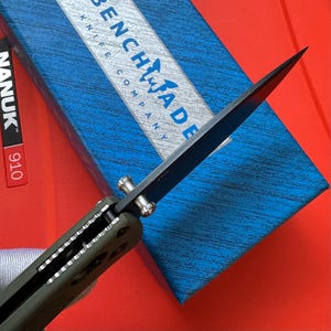 May include: A close-up of a Benchmade knife with a black blade and olive green handle. The knife is partially open, revealing the blade's sharp edge. The Benchmade logo is visible on a blue and white box in the background. The knife is set against a red surface.