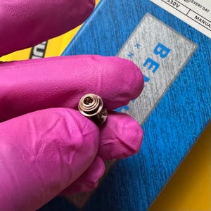 May include: A small, metallic, cylindrical component with a recessed center and a tiered, circular top edge. The object is held between fingers covered in bright pink gloves, positioned against a blue box with partial text reading BE.