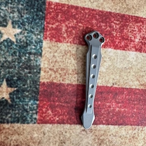 Pu&ograve; includere: Un utensile in metallo color argento con una serie di fori circolari e una forma unica. L'utensile presenta un design a tre fori nella parte superiore e un'estremit&agrave; appuntita. Lo sfondo &egrave; una bandiera americana.