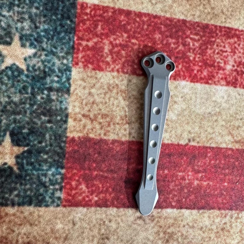 Pu&ograve; includere: Un utensile in metallo color argento con una serie di fori circolari e una forma unica. L'utensile presenta un design a tre fori nella parte superiore e un'estremit&agrave; appuntita. Lo sfondo &egrave; una bandiera americana.