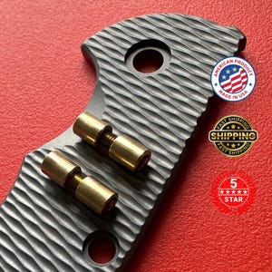 May include: Close-up of a textured, dark gray metal object with two gold-colored cylindrical components. The object has a circular hole and is marked with "AMERICAN PRODUCT MADE IN USA".