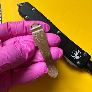 May include: A metallic bronze pocket knife clip featuring an engraved koi fish pattern, positioned over a black handle of an out-the-front knife. The clip has three small circular mounting holes at the top and a tapered shape with a curved end.