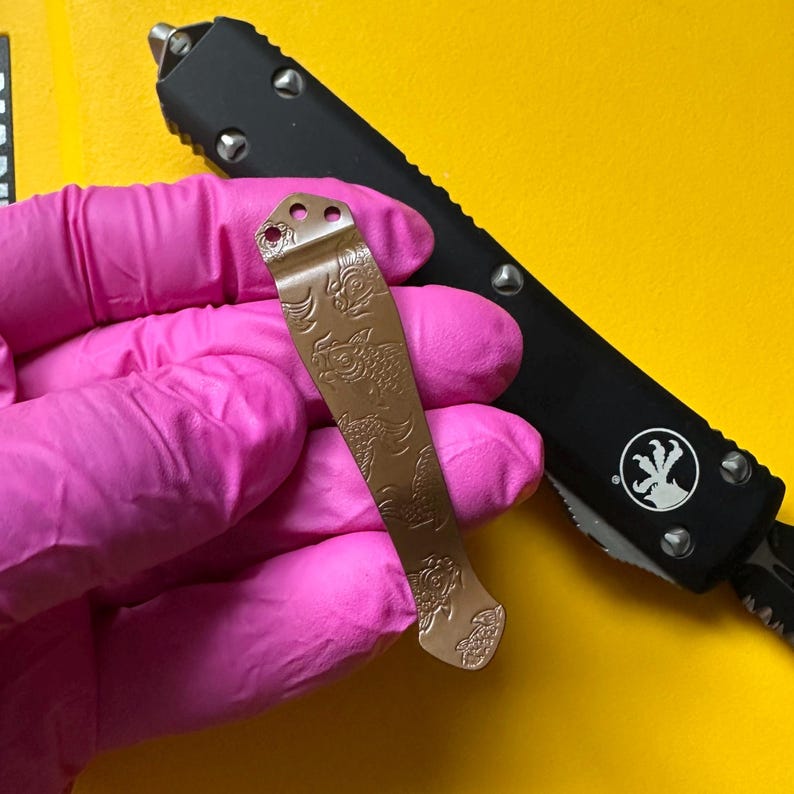 May include: A metallic bronze pocket knife clip featuring an engraved koi fish pattern, positioned over a black handle of an out-the-front knife. The clip has three small circular mounting holes at the top and a tapered shape with a curved end.