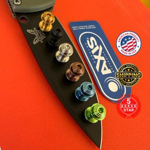 May include: A black Benchmade knife blade with a variety of colored thumb studs. The studs are gold, black, silver, copper, blue, and green. A blue and white Axis Mechanics card and an American product sticker are also present.