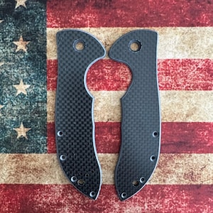 May include: Two black carbon fiber knife scales with a checkered pattern. Each scale has a curved shape with a hole at the top and several smaller holes along the edge. The scales are displayed against a backdrop of the American flag.
