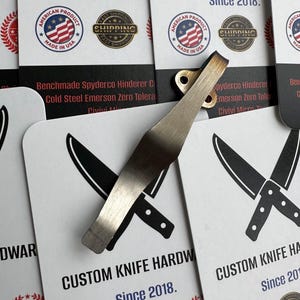 May include: A close-up of a custom knife hardware product. The image shows a silver pocket clip with a brass rivet. The clip is over white cards with black knife illustrations and text: "CUSTOM KNIFE HARDWARE Since 2018."