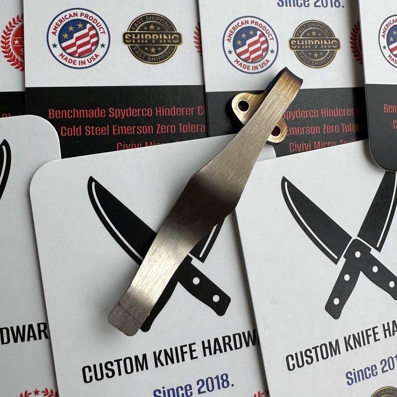 May include: A close-up of a custom knife hardware product. The image shows a silver pocket clip with a brass rivet. The clip is over white cards with black knife illustrations and text: "CUSTOM KNIFE HARDWARE Since 2018."