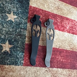 May include: Two metal pocket clips, one black and one silver, each featuring a cut-out Spartan helmet design. The clips are displayed against a backdrop of the American flag, showcasing a patriotic theme.