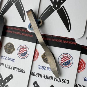 Op de afbeelding: Een op maat gemaakt mes hardware-onderdeel met een gebogen, metalen ontwerp. De hardware heeft een licht zilveren kleur met hints van blauw en paars. De achtergrond bevat kaarten met de tekst "CUSTOM KNIFE HARDWARE" en "MADE IN USA."