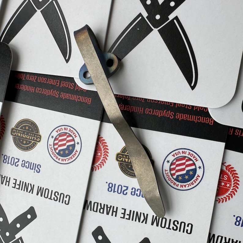 Op de afbeelding: Een op maat gemaakt mes hardware-onderdeel met een gebogen, metalen ontwerp. De hardware heeft een licht zilveren kleur met hints van blauw en paars. De achtergrond bevat kaarten met de tekst "CUSTOM KNIFE HARDWARE" en "MADE IN USA."