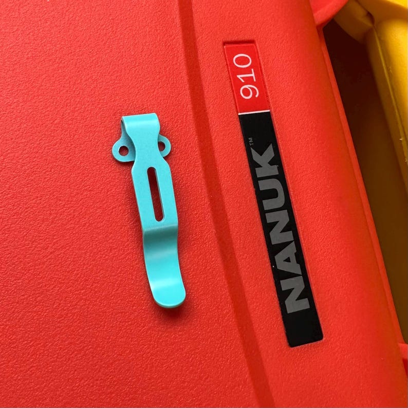 May include: A close-up of a teal-colored clip on a red surface. The clip is made of metal and has a curved design with a slot in the middle. The red surface has the text "NANUK 910".
