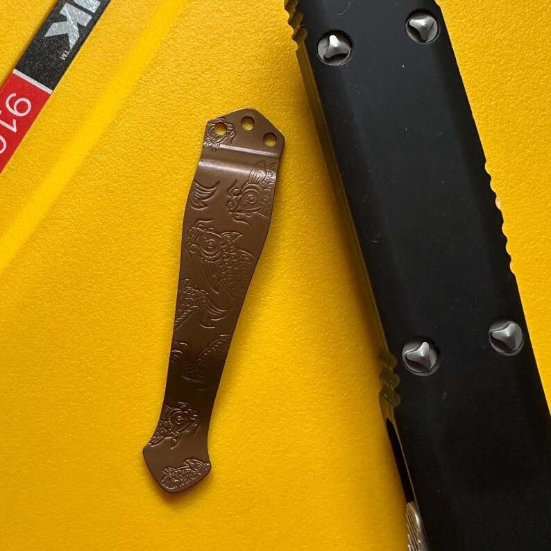 May include: A brown metal pocket knife clip featuring an engraved koi fish pattern, positioned next to a black handle of a folding knife. The clip has three circular mounting holes at the top and a curved shape, displaying detailed fish motifs.