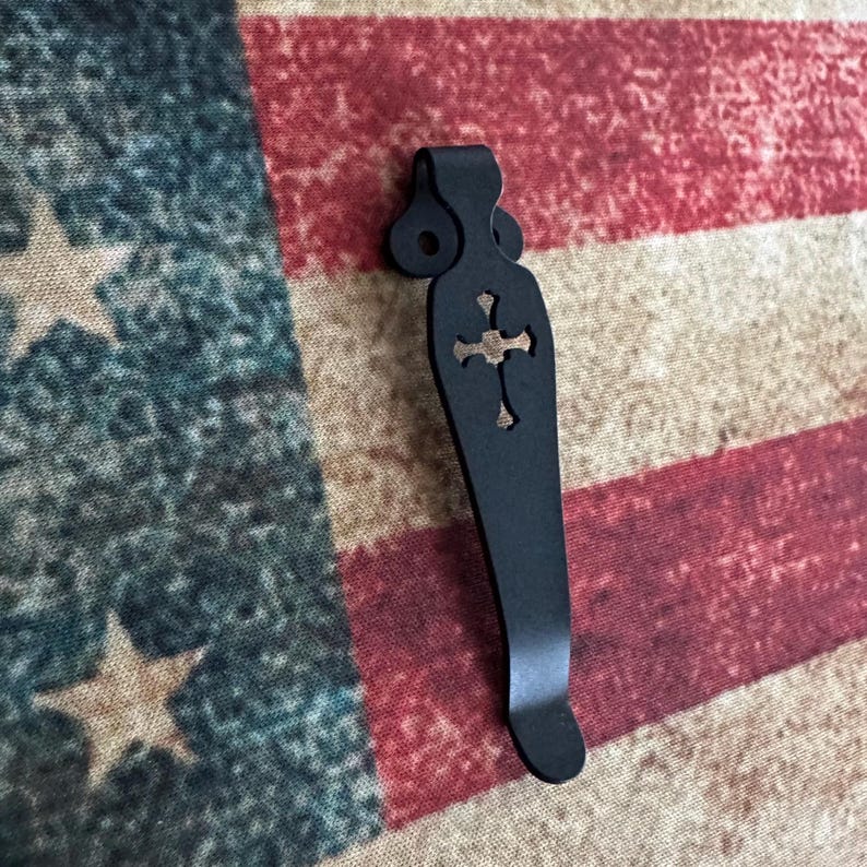 K&ouml;nnte beinhalten: Ein schwarzer Metall-Taschenclip mit einem Kreuz-Ausschnitt-Design. Der Clip ist gebogen und hat oben eine Schlaufe zur Befestigung. Der Hintergrund ist eine verblasste amerikanische Flagge.