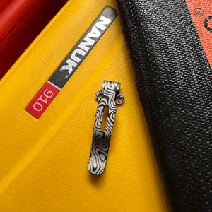 K&ouml;nnte beinhalten: Ein gemusterter Metallclip mit einem Wirbeldesign in Schwarz und Silber, der auf einer gelben Oberfl&auml;che liegt. Der Clip befindet sich neben einem schwarzen strukturierten Objekt und einem Etikett mit der Aufschrift "NANUK 910".