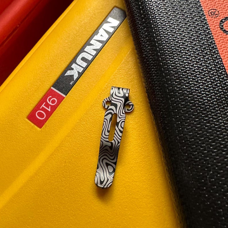 K&ouml;nnte beinhalten: Ein gemusterter Metallclip mit einem Wirbeldesign in Schwarz und Silber, der auf einer gelben Oberfl&auml;che liegt. Der Clip befindet sich neben einem schwarzen strukturierten Objekt und einem Etikett mit der Aufschrift "NANUK 910".