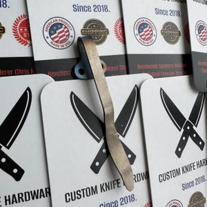 Op de afbeelding: Een close-up van een op maat gemaakte hardwareclip voor messen met een metallic afwerking. De clip is geplaatst over witte kaarten met gekruiste mesillustraties en de tekst "CUSTOM KNIFE HARDWARE Since 2018". De kaarten tonen ook "AMERICAN PRODUCT MADE IN USA" zegels.
