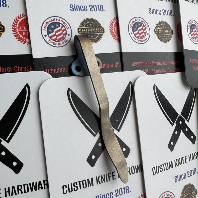 Op de afbeelding: Een close-up van een op maat gemaakte hardwareclip voor messen met een metallic afwerking. De clip is geplaatst over witte kaarten met gekruiste mesillustraties en de tekst "CUSTOM KNIFE HARDWARE Since 2018". De kaarten tonen ook "AMERICAN PRODUCT MADE IN USA" zegels.