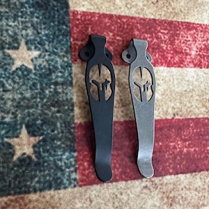 May include: Two metal pocket clips, one black and one silver, each featuring a Spartan helmet design cut-out. The clips are displayed against a backdrop of a faded American flag, showcasing a patriotic theme.