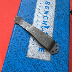 Op de afbeelding: Een zilverkleurige mesclip met een gegraveerd koi-visontwerp. De clip is gebogen en heeft aan &eacute;&eacute;n uiteinde drie kleine gaatjes. Het woord "BENCHMADE" is zichtbaar op een blauwe doos op de achtergrond.