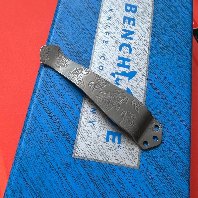 Op de afbeelding: Een zilverkleurige mesclip met een gegraveerd koi-visontwerp. De clip is gebogen en heeft aan &eacute;&eacute;n uiteinde drie kleine gaatjes. Het woord "BENCHMADE" is zichtbaar op een blauwe doos op de achtergrond.