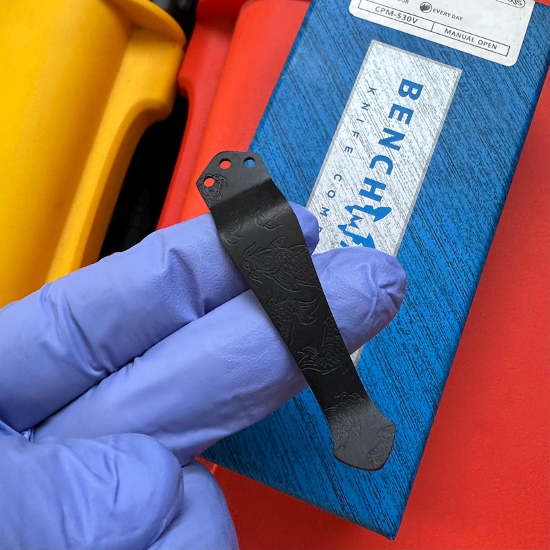 Op de afbeelding: Een zwarte metalen mesclip met een gegraveerd visschubbenpatroon. De clip heeft drie gaten aan de bovenkant en wordt in een gehandschoende hand gehouden. De Benchmade Knife Co. doos staat op de achtergrond.