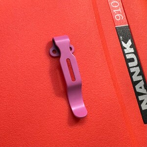 May include: A pink metal clip with a curved design and a rectangular cutout. The clip is set against a textured red background. The word "NANUK" is visible on a black and red label.