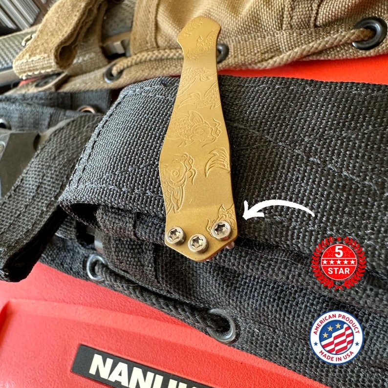 Puede incluir: Un clip met&aacute;lico dorado con un dise&ntilde;o floral grabado, sujeto a una tela negra. El clip se asegura con tres tornillos. La imagen tambi&eacute;n incluye una insignia "5 STAR" roja y blanca y una insignia "American Product Made in USA".
