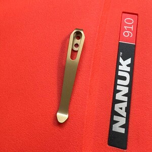 May include: A gold-colored metal pocket clip with two holes and a curved end. The clip is next to a red and black label with the text "NANUK 910".