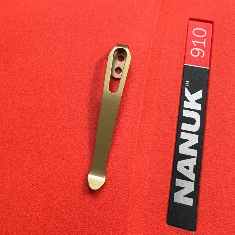 May include: A gold-colored metal pocket clip with two holes and a curved end. The clip is next to a red and black label with the text "NANUK 910".