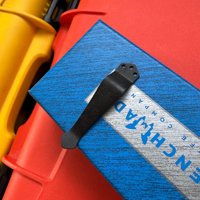 Op de afbeelding: Een zwarte metalen zakclip met een decoratief patroon, rustend op een blauwe doos met het woord "BENCHMADE" zichtbaar. De clip heeft drie gaten aan de bovenkant en een gebogen ontwerp.