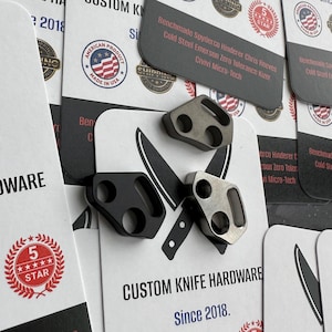 May include: Close-up of custom knife hardware, including three metal pieces in black and silver. The pieces have circular cutouts and are displayed on cards with text that reads "CUSTOM KNIFE HARDWARE Since 2018."
