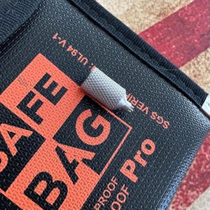 May include: A close-up of a silver metal tool with a textured grip, resting on a black bag with orange text that reads "SAFE BAG Pro". The bag also has the text "UL94 V-1" and "SGS VERIFIED".