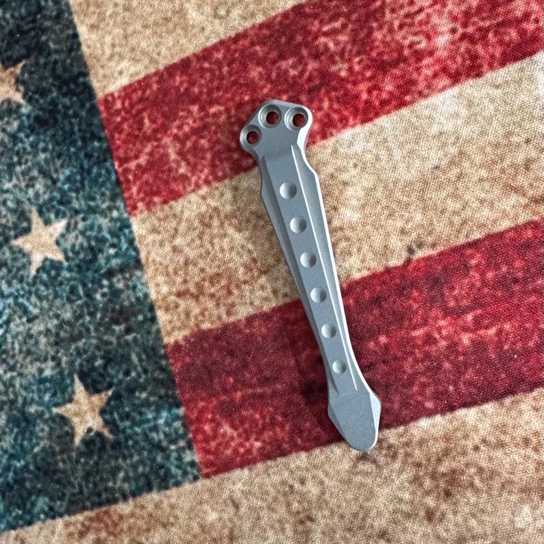 Pu&ograve; includere: Un utensile in metallo color argento con una serie di rientranze circolari e tre fori nella parte superiore. L'utensile &egrave; posto su uno sfondo di una bandiera americana rossa, bianca e blu.