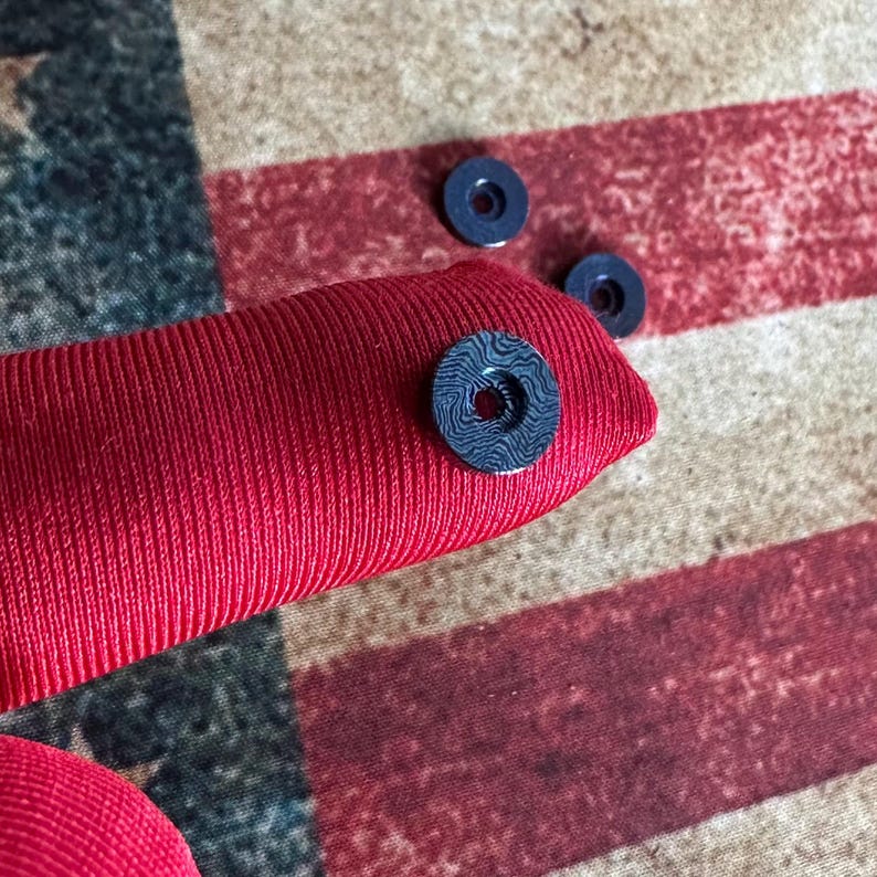 Pu&ograve; includere: Primo piano di un oggetto in tessuto rosso con tre piccole rondelle blu scuro. Una rondella &egrave; attaccata al tessuto e presenta un motivo a vortice unico. Lo sfondo &egrave; una bandiera americana sbiadita.