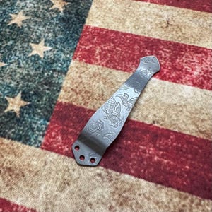 May include: A silver-colored metal knife clip with an engraved koi fish design. The clip has three holes at the base for attachment. The background is an American flag.