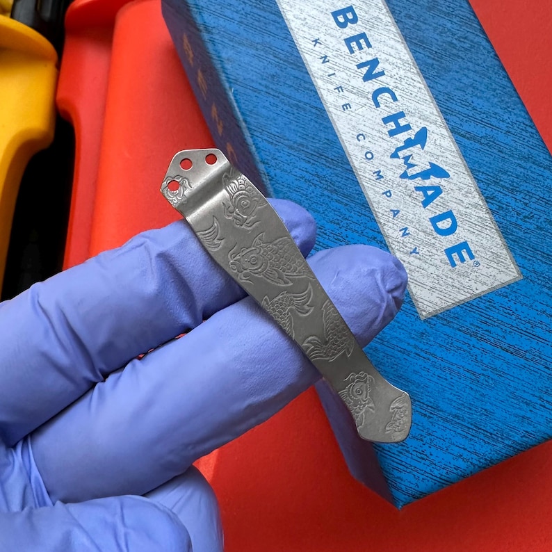 Op de afbeelding: Een zilverkleurige mesclip met een gegraveerd vispatroon. De clip heeft drie gaten aan de bovenkant en een gebogen ontwerp. De doos van Benchmade Knife Company is zichtbaar op de achtergrond.