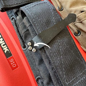 May include: Close-up of a black metal clip attached to a black fabric strap with silver screws. The clip has a decorative pattern. The background includes a red surface with the text "NANUK 910" and a tan fabric.