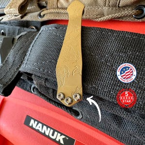 May include: A gold-colored metal clip with an engraved floral design, attached to a black fabric. The clip is secured with three silver screws. The image also includes a red case with the text "NANUK" and a "Made in USA" sticker.