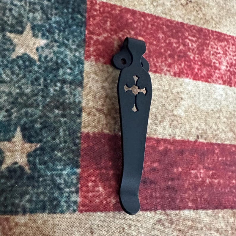 K&ouml;nnte beinhalten: Ein schwarzer Metall-Taschenclip mit einem dekorativen Kreuz-Ausschnitt. Der Clip hat ein gebogenes Design und eine Schlaufe oben. Der Hintergrund ist eine amerikanische Flagge.