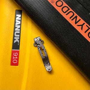 K&ouml;nnte beinhalten: Ein gemusterter Metallclip mit einem wellenf&ouml;rmigen Design, der auf einer gelben Oberfl&auml;che liegt. Der Clip befindet sich neben einem schwarzen Objekt mit orangefarbenem Text und einem schwarz-roten Etikett mit der Aufschrift "NANUK 910".
