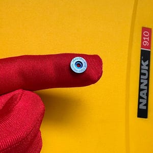以下が含まれることがあります： 赤い布のクローズアップ。小さな丸い青と銀色のボタンには「AUTOMATIC COLT PISTOL」と書かれています。背景は明るい黄色の表面で、「NANUK 910」の文字があります。