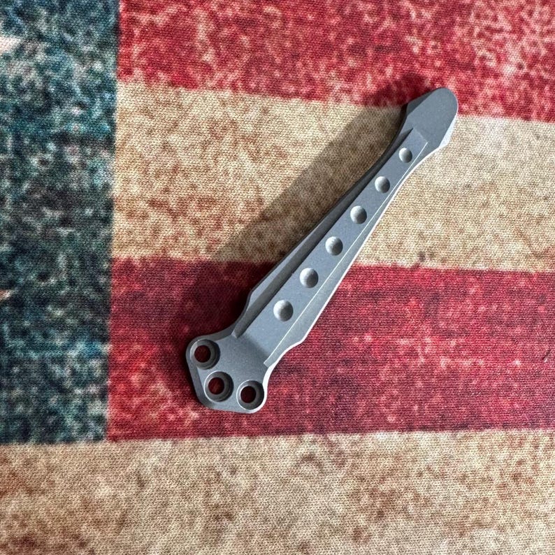Pu&ograve; includere: Un utensile in metallo color argento con una serie di fori circolari e una forma unica. L'utensile &egrave; esposto su uno sfondo di una bandiera americana rossa, bianca e blu.