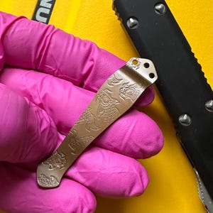 May include: A metallic pocket knife clip featuring an engraved koi fish pattern. The clip has a bronze-toned finish with a curved shape and two circular mounting holes at the top end, positioned next to a black handle with silver hardware.