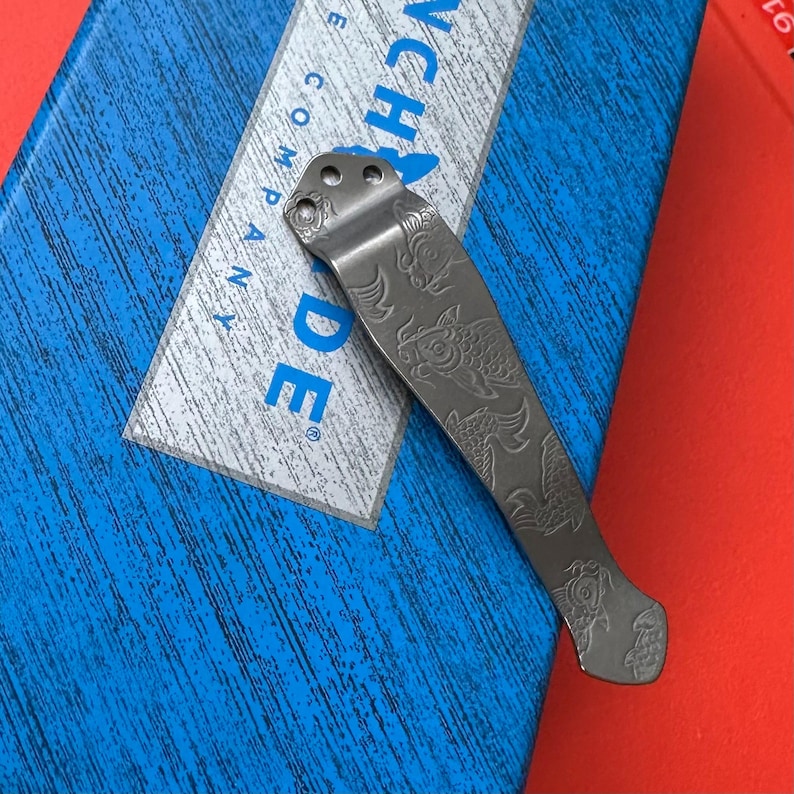 Op de afbeelding: Een zilverkleurige metalen zakclip met een gegraveerd koi-visontwerp. De clip heeft drie gaten aan de bovenkant voor bevestiging en ligt op een blauw en wit oppervlak met de tekst "Benchmade Company".