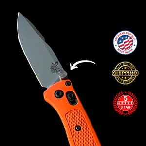 K&ouml;nnte beinhalten: Ein orangefarbenes Klappmesser mit silberner Klinge. Das Messer hat einen strukturierten Griff und ein Schmetterlingslogo auf der Klinge. Das Bild enth&auml;lt ein "Made in USA"-Abzeichen und ein "5 Sterne"-Abzeichen.